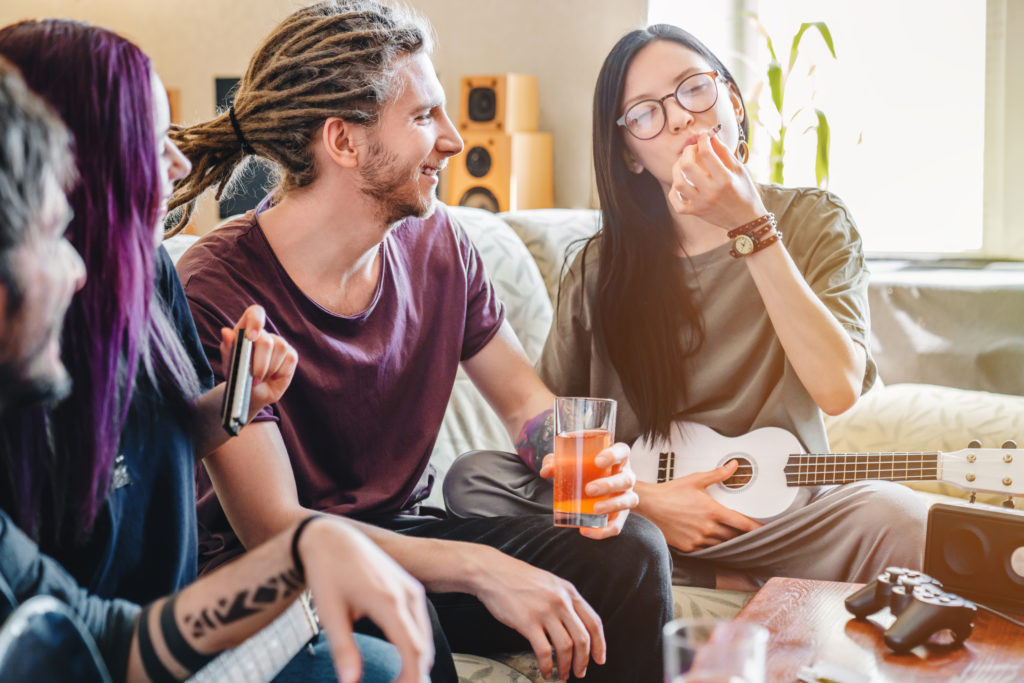Young female smoking joint with cannabis while playing on music instruments at home with friends